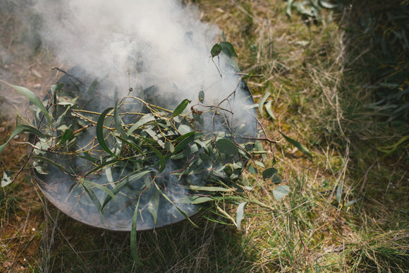 A metal bowl being used in a smoking ceremony sits on grass. It has been filled with branches of native Australian trees such as eucalyptus, which are smouldering above the fire beneath them.