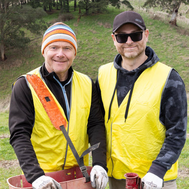 Two men in yellow vests standing in a grassy field with tools, surrounded by other people in similar attire.