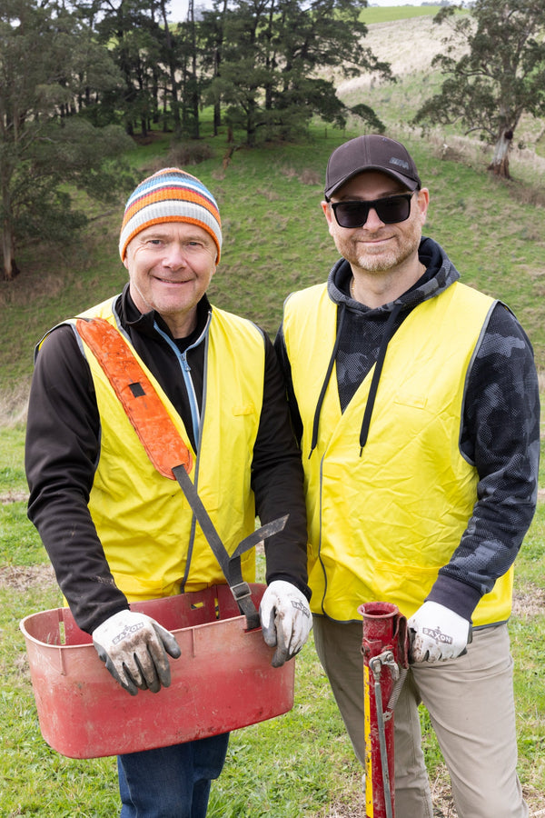 Two men in yellow vests standing in a grassy field with tools, surrounded by other people in similar attire.