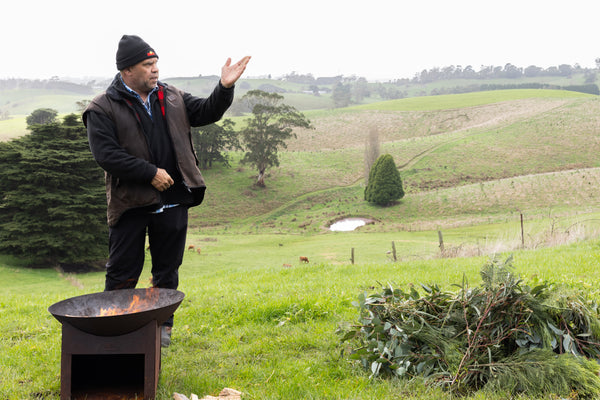 Boonwurrung man Willie Pepper stands in a rolling green landscape as he welcomes guests to his Country with a small fire in a metal bowl burning beside him. 